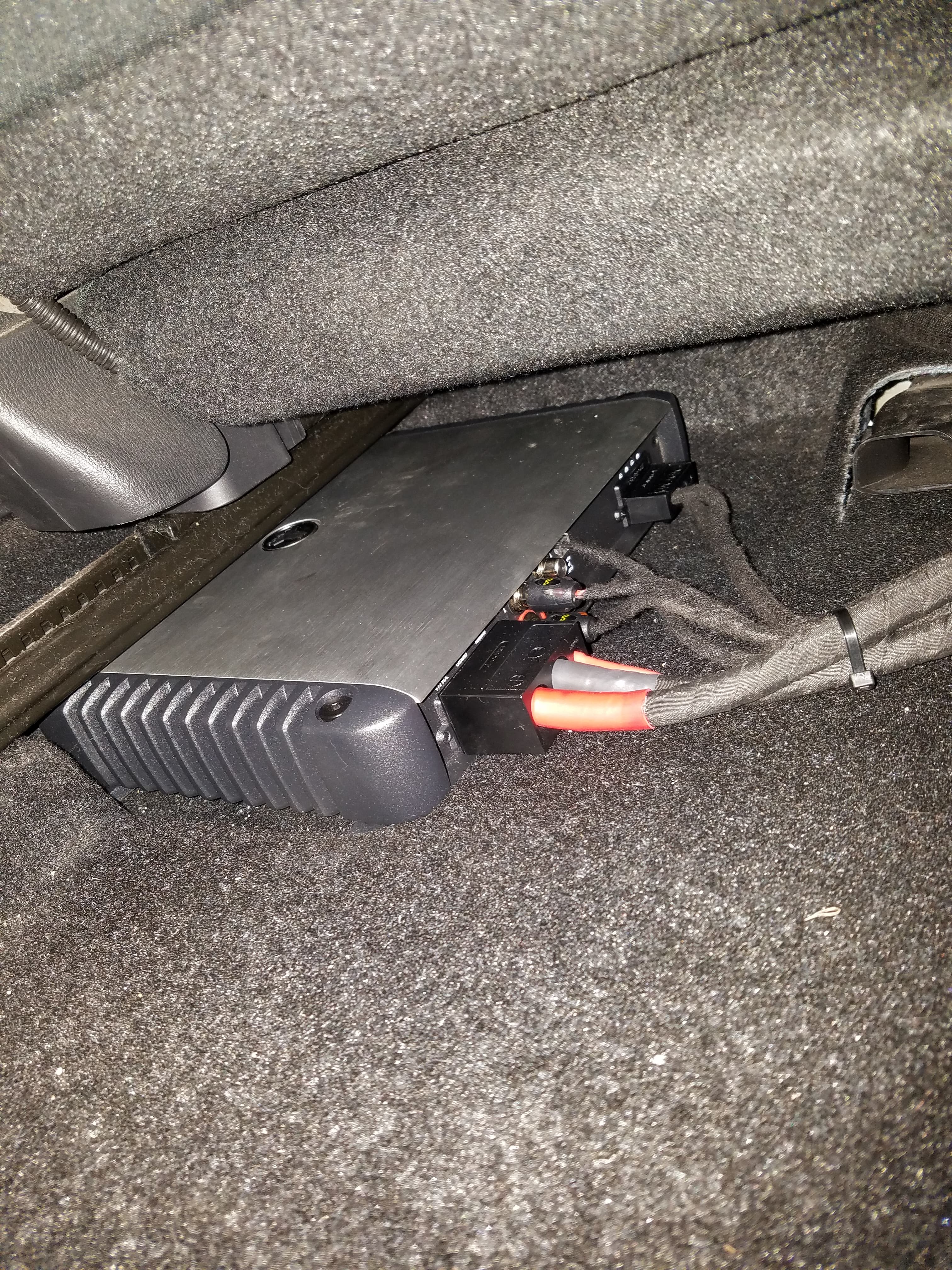 Silver car amplifier with multiple wires connected, installed on carpet under a vehicle seat.