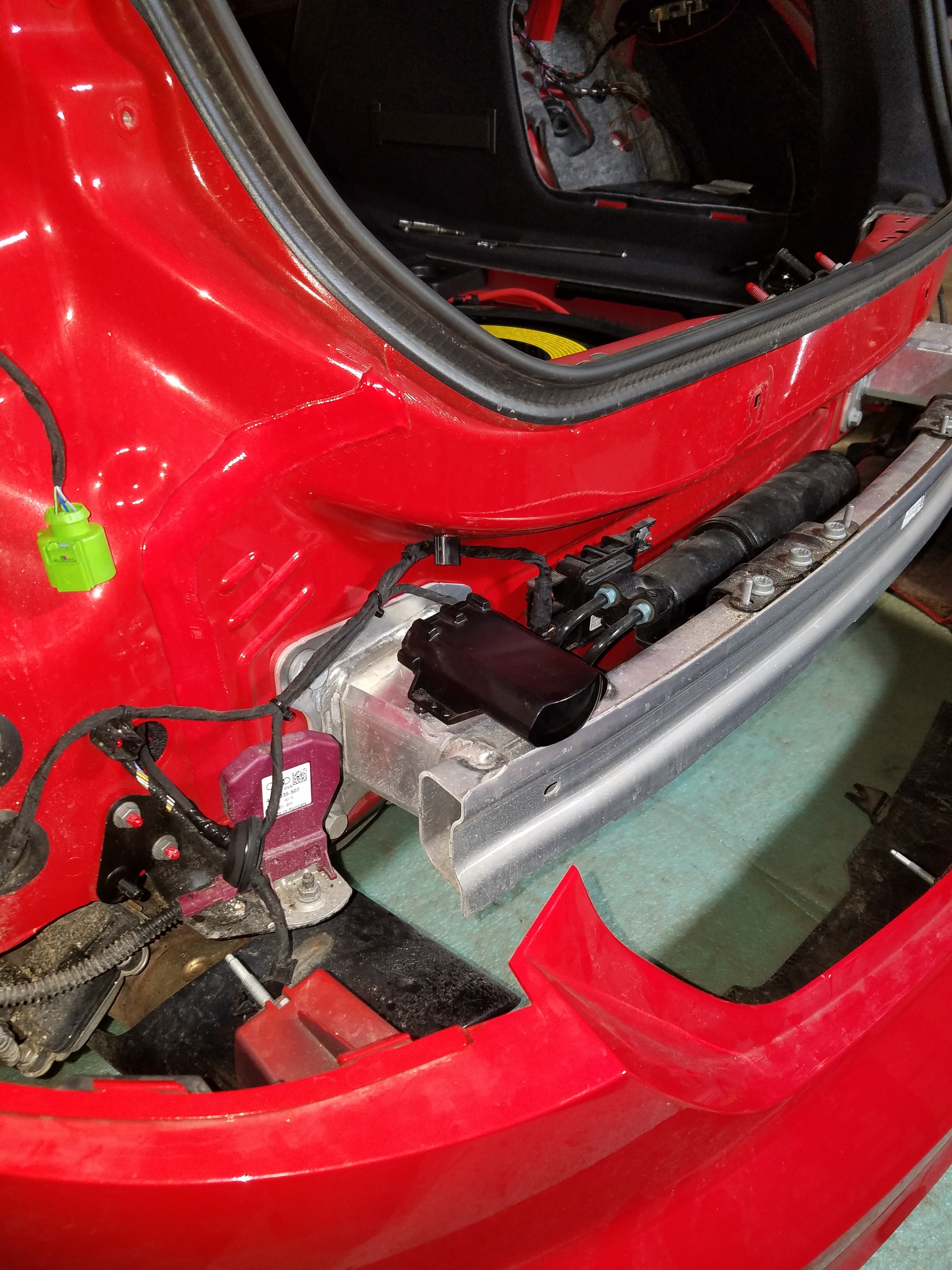 Exposed rear of a red car showing silver bumper bar, black sensors, and electrical components.