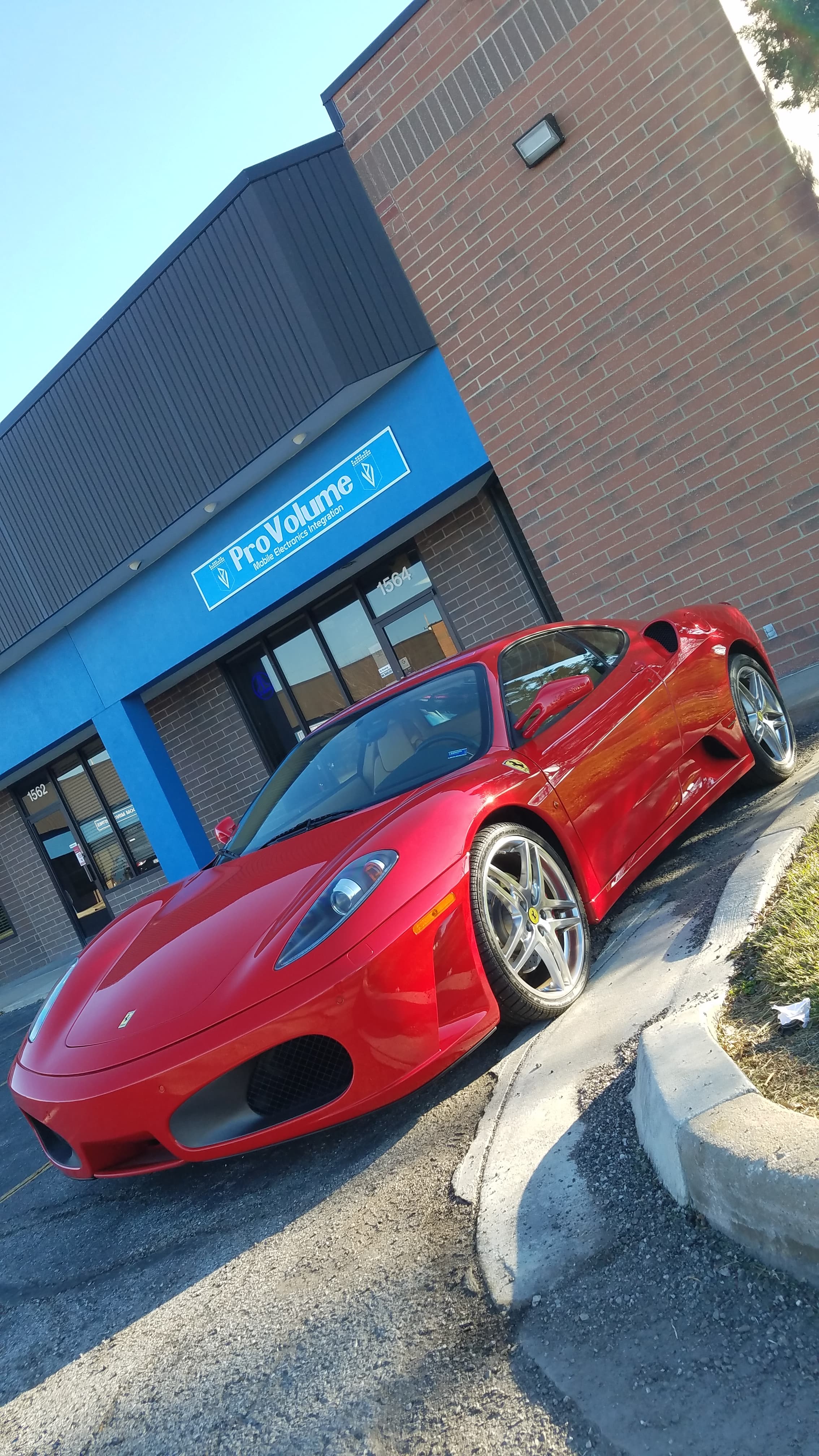 Red Ferrari F430 parked outside a ProVolume electronics store on a bright, sunny day.