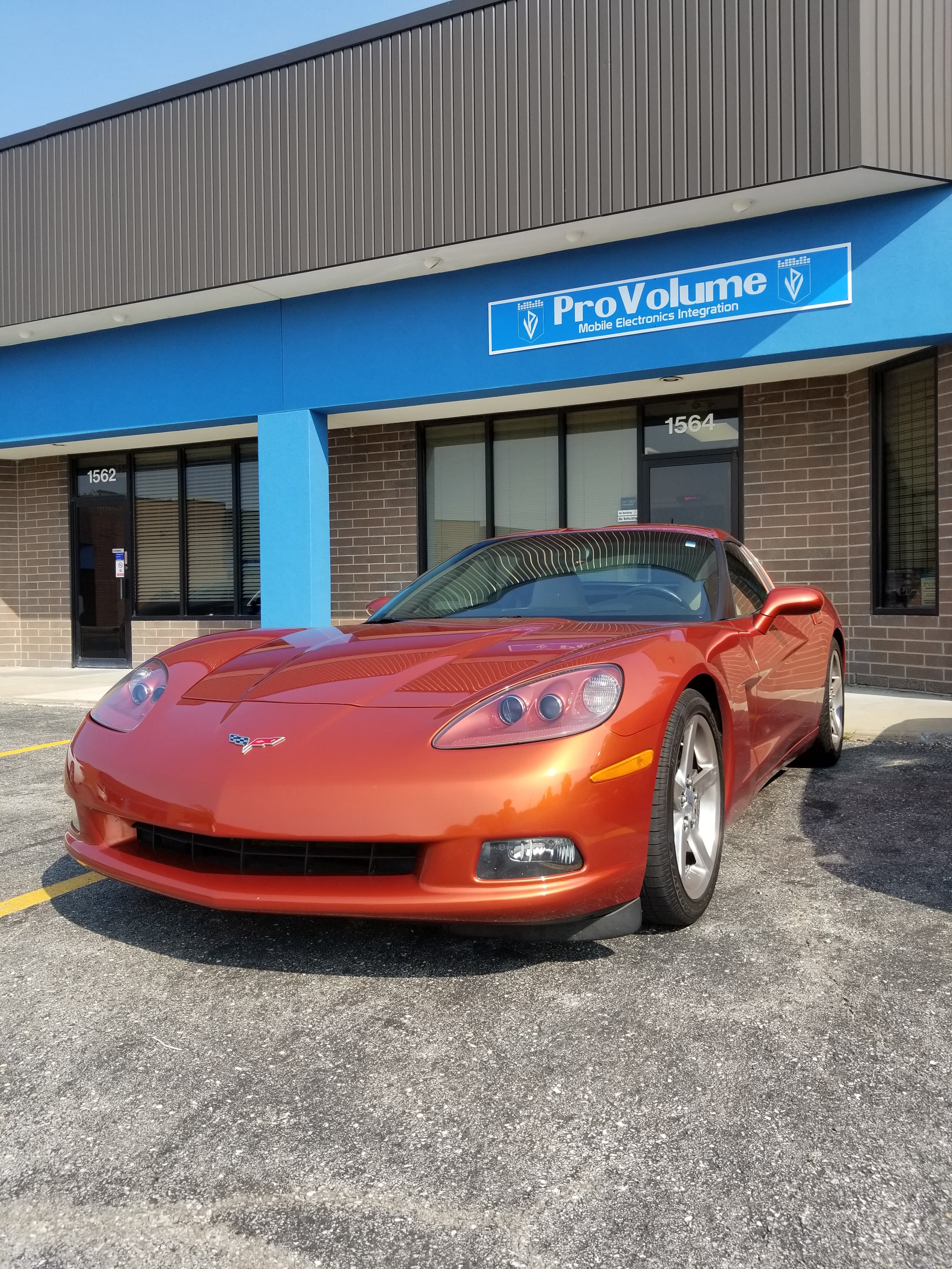 A shiny orange Chevrolet Corvette parked in front of a ProVolume mobile electronics integration store.