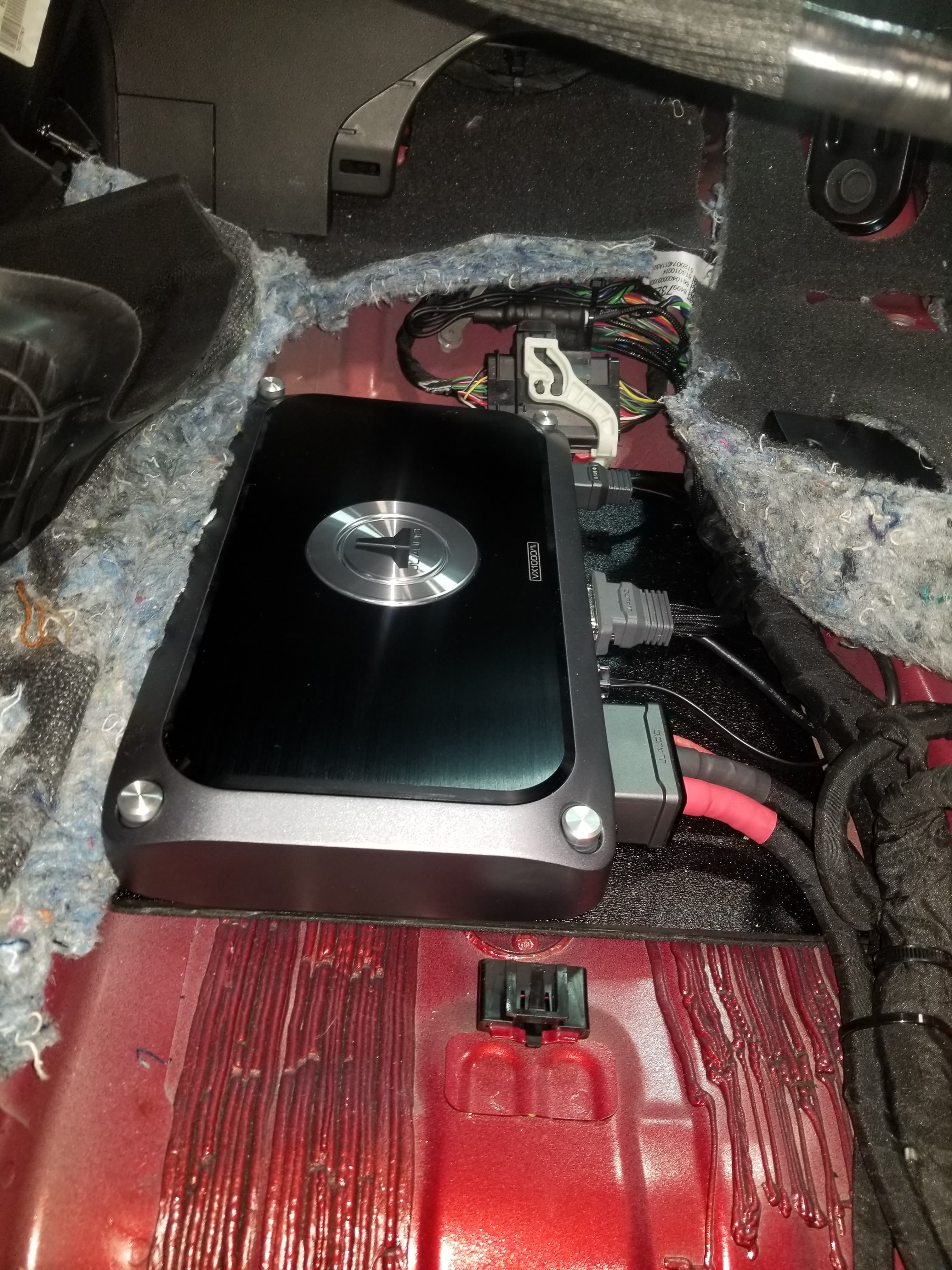 Black JL Audio amplifier installed in a vehicle's floorboard with exposed wiring and grey insulation.