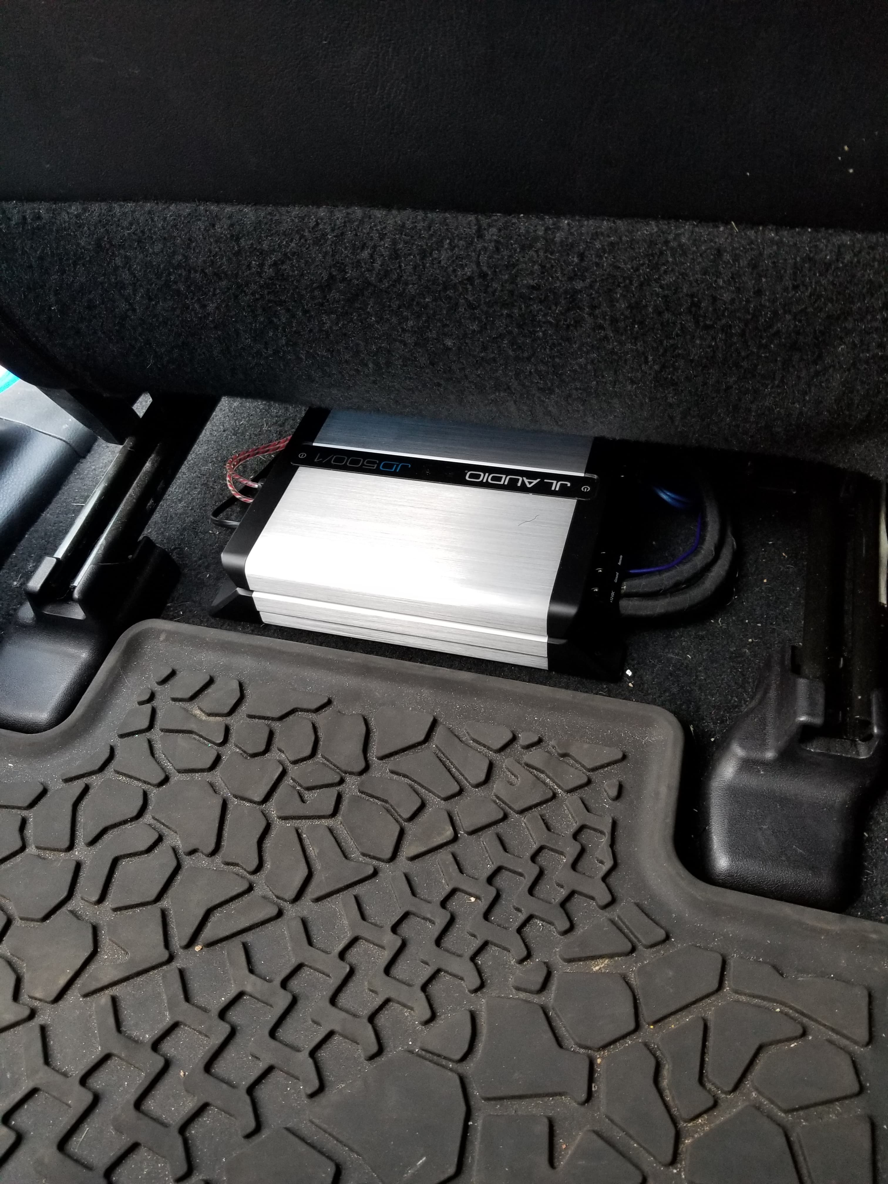Silver JL Audio JD500/1 amplifier installed under a car seat beside a rubber mat.