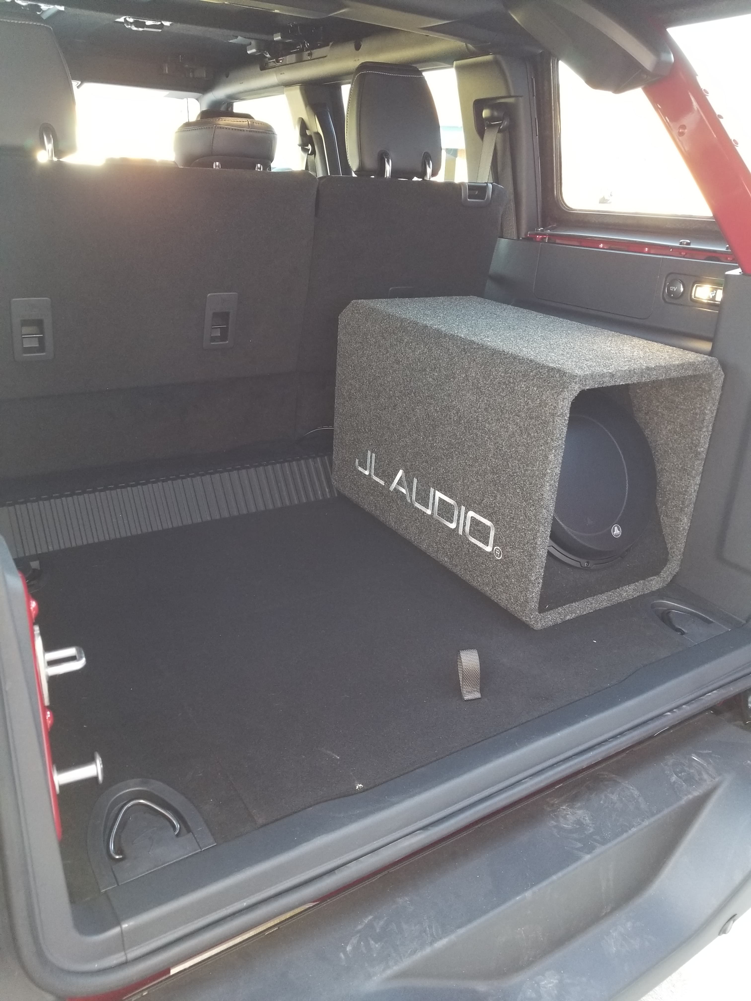 JL Audio subwoofer in a grey carpeted box inside a black vehicle cargo area.