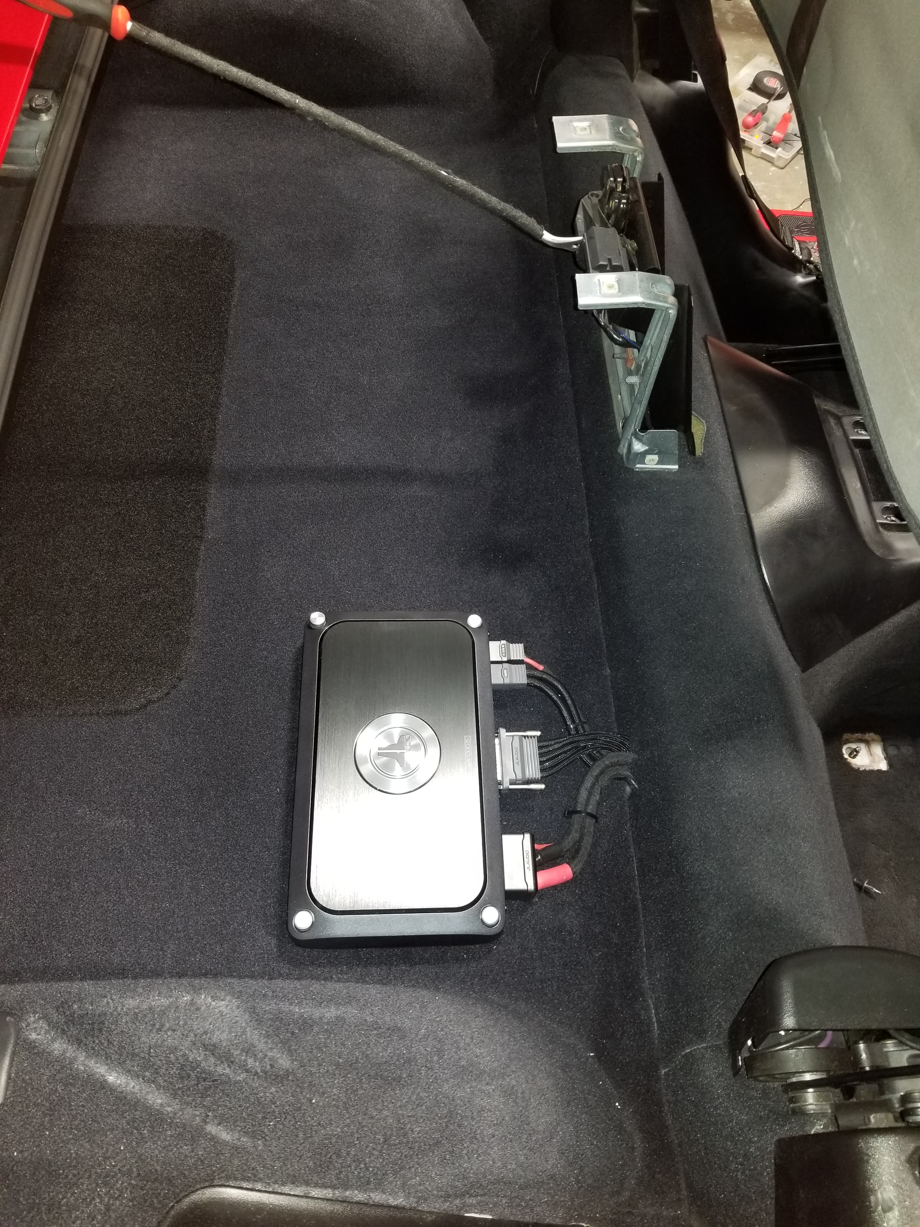 Black JL Audio amplifier mounted on carpeted vehicle interior with connected wiring and cables.