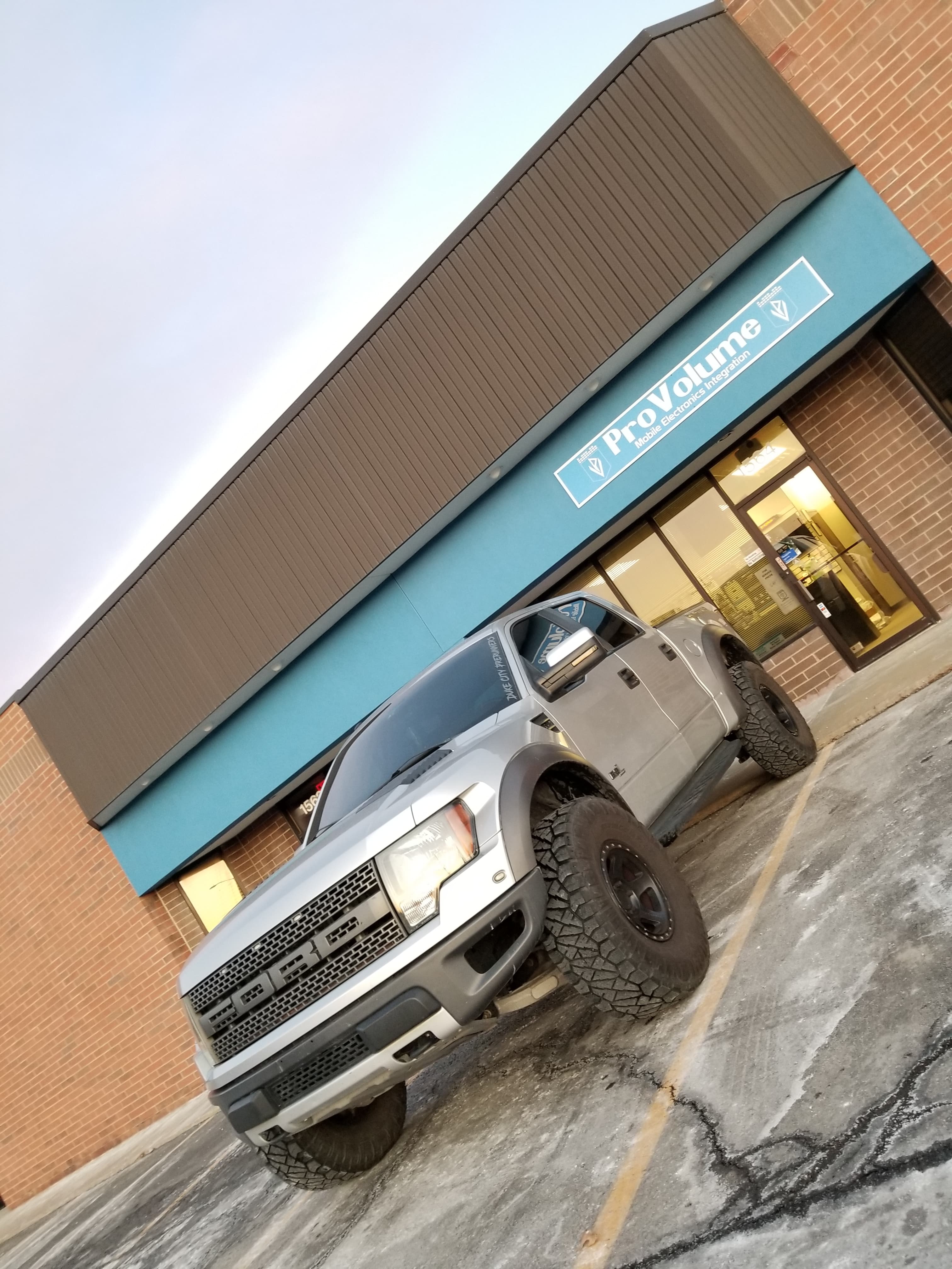 Silver Ford Raptor truck with large tires parked in front of a ProVolume electronics store.