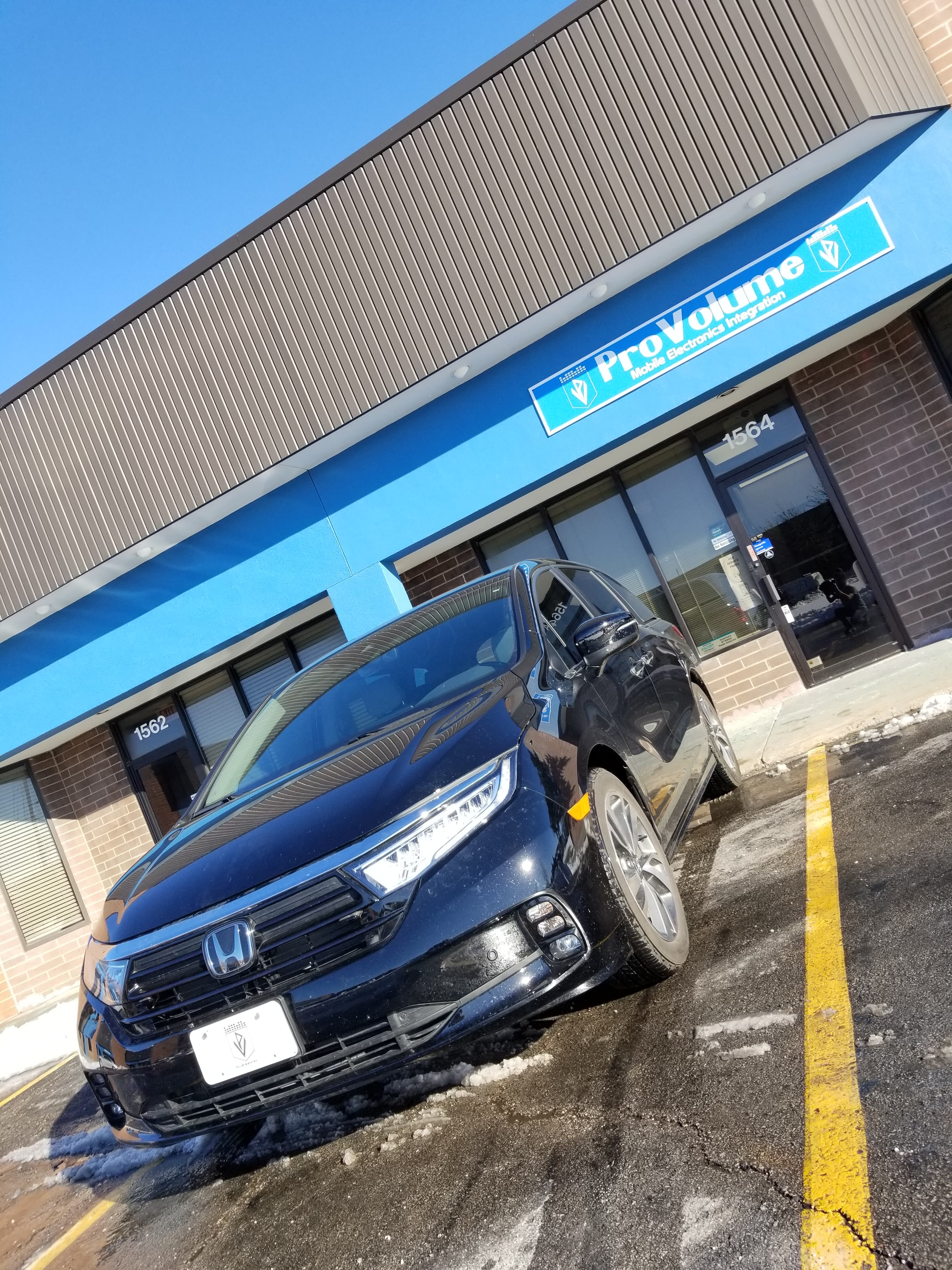 Black Honda minivan parked outside a ProVolume Mobile Electronics store under a clear blue sky.