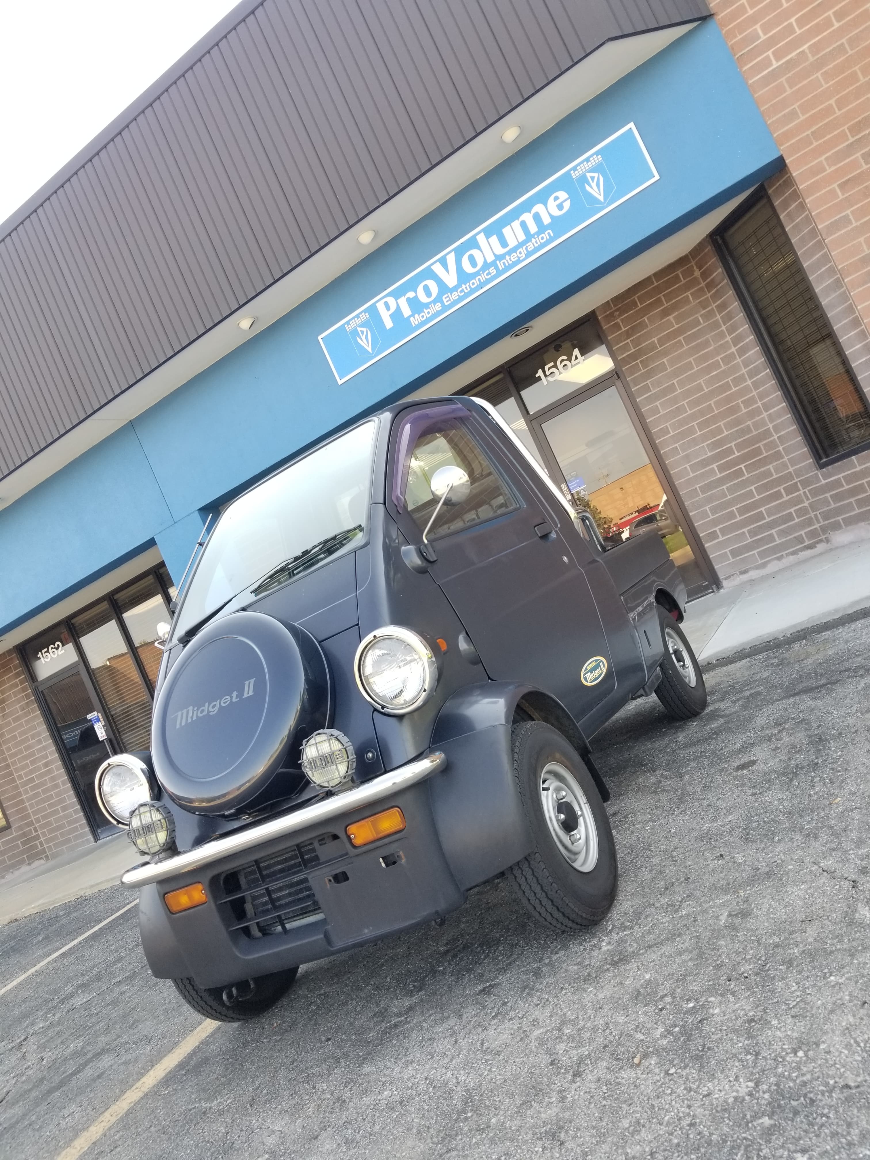 Dark blue Daihatsu Midget II micro-truck parked in front of a ProVolume electronics store.