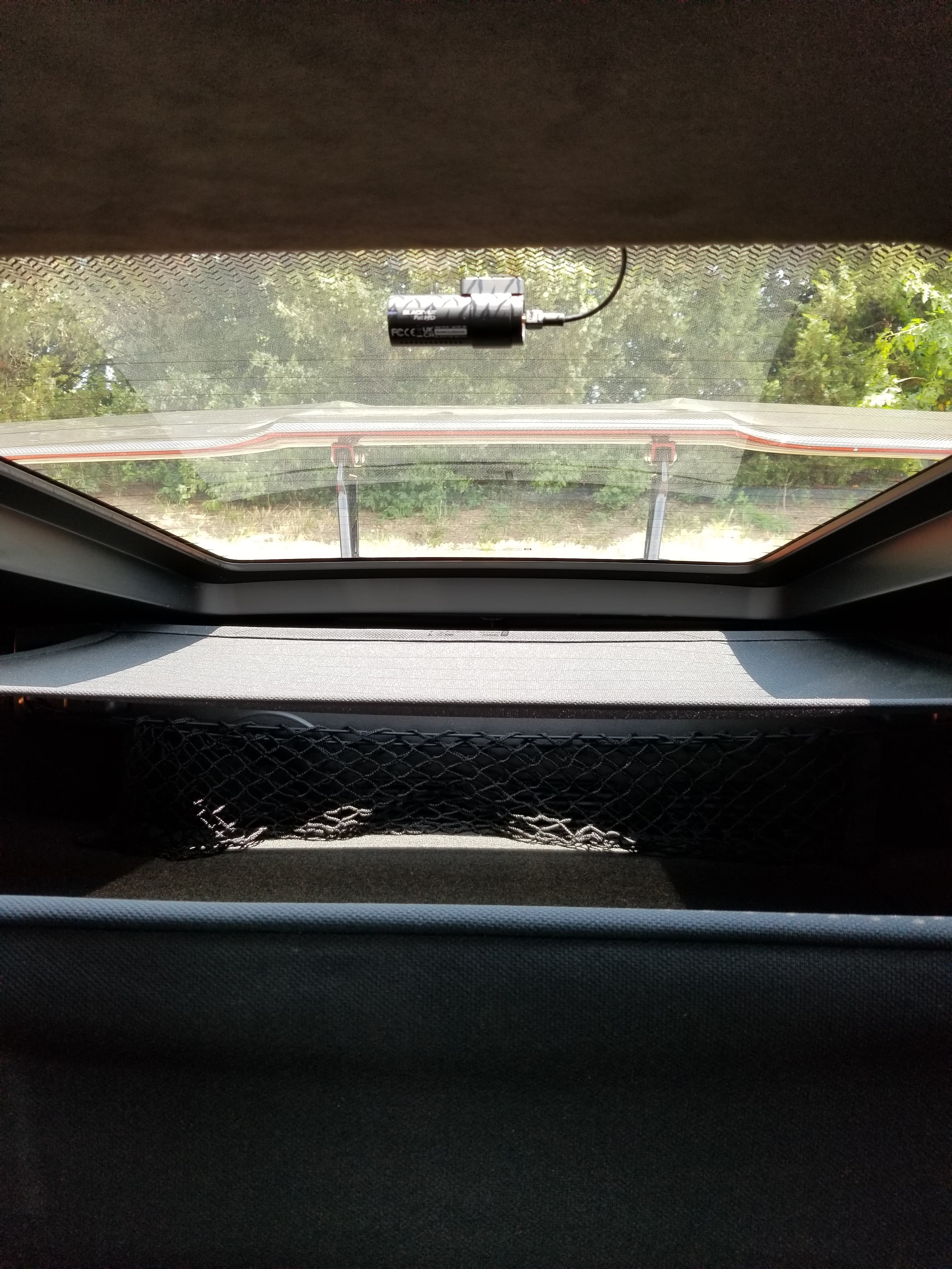 Rear dashcam mounted on a car window above a cargo net, overlooking trees outside.