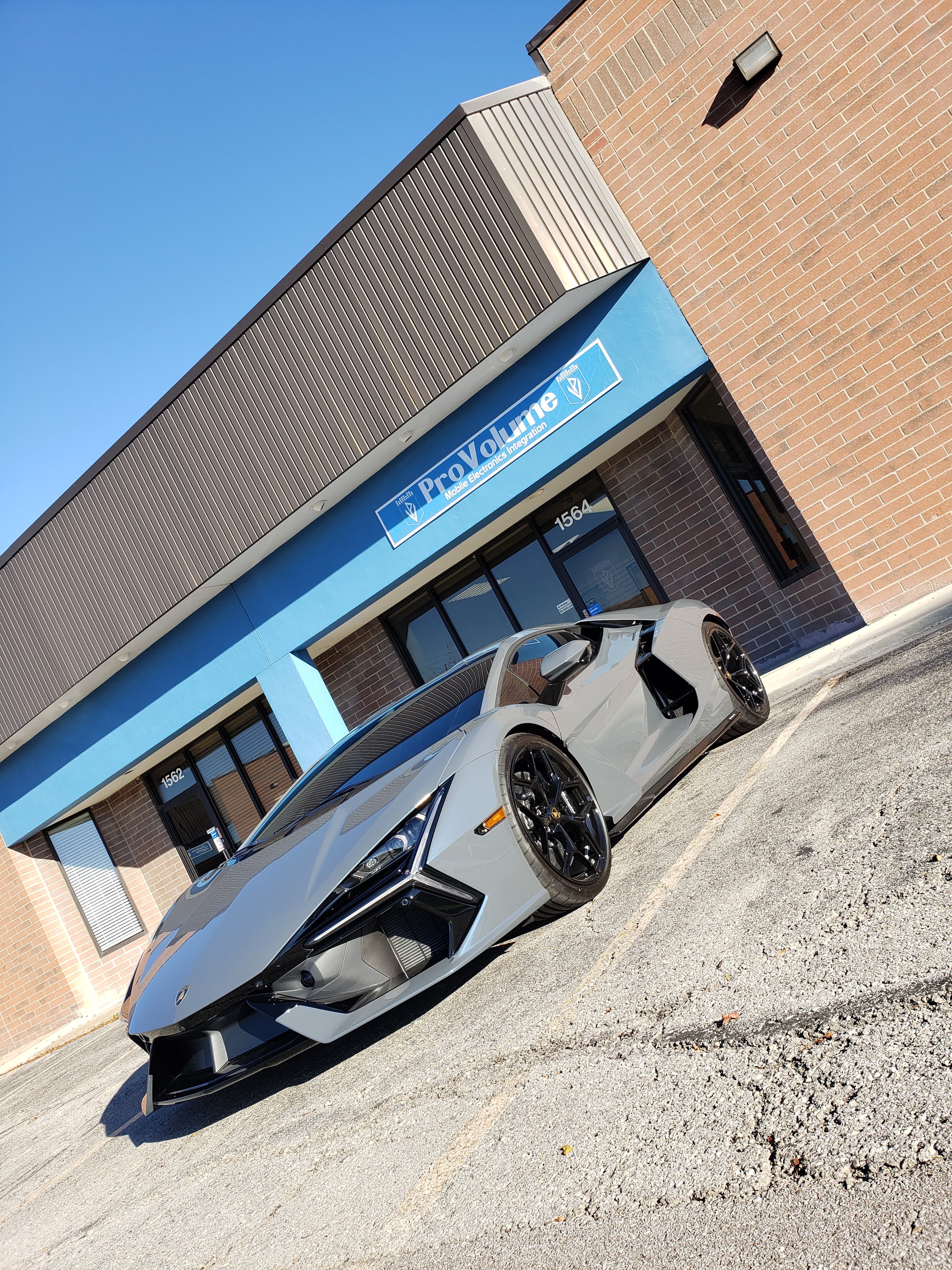 Sleek grey Lamborghini Revuelto parked outside a ProVolume electronics store on a sunny day.