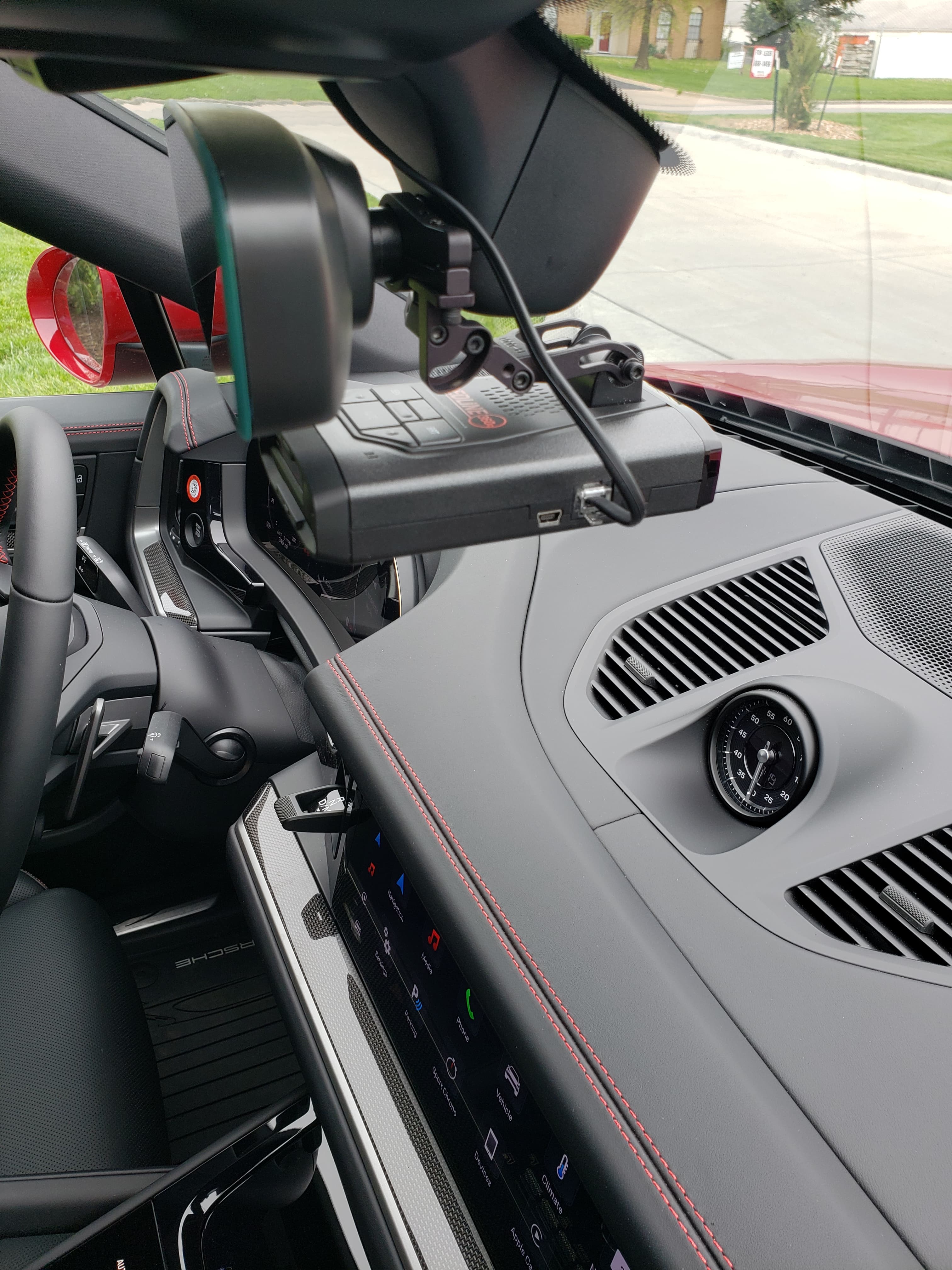 Radar detector mounted to the rearview mirror of a Porsche with red-stitched leather dashboard.