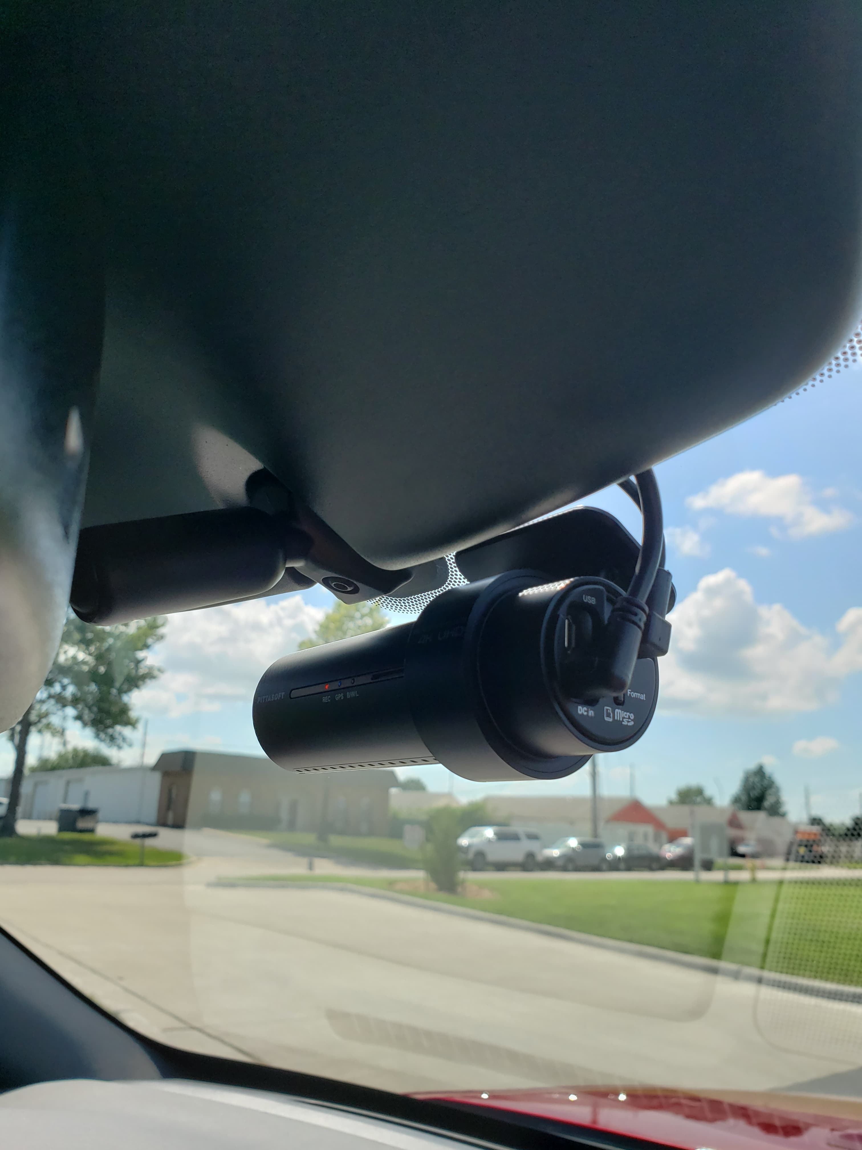 Black dashcam mounted behind a car's rearview mirror, overlooking a sunny parking lot.