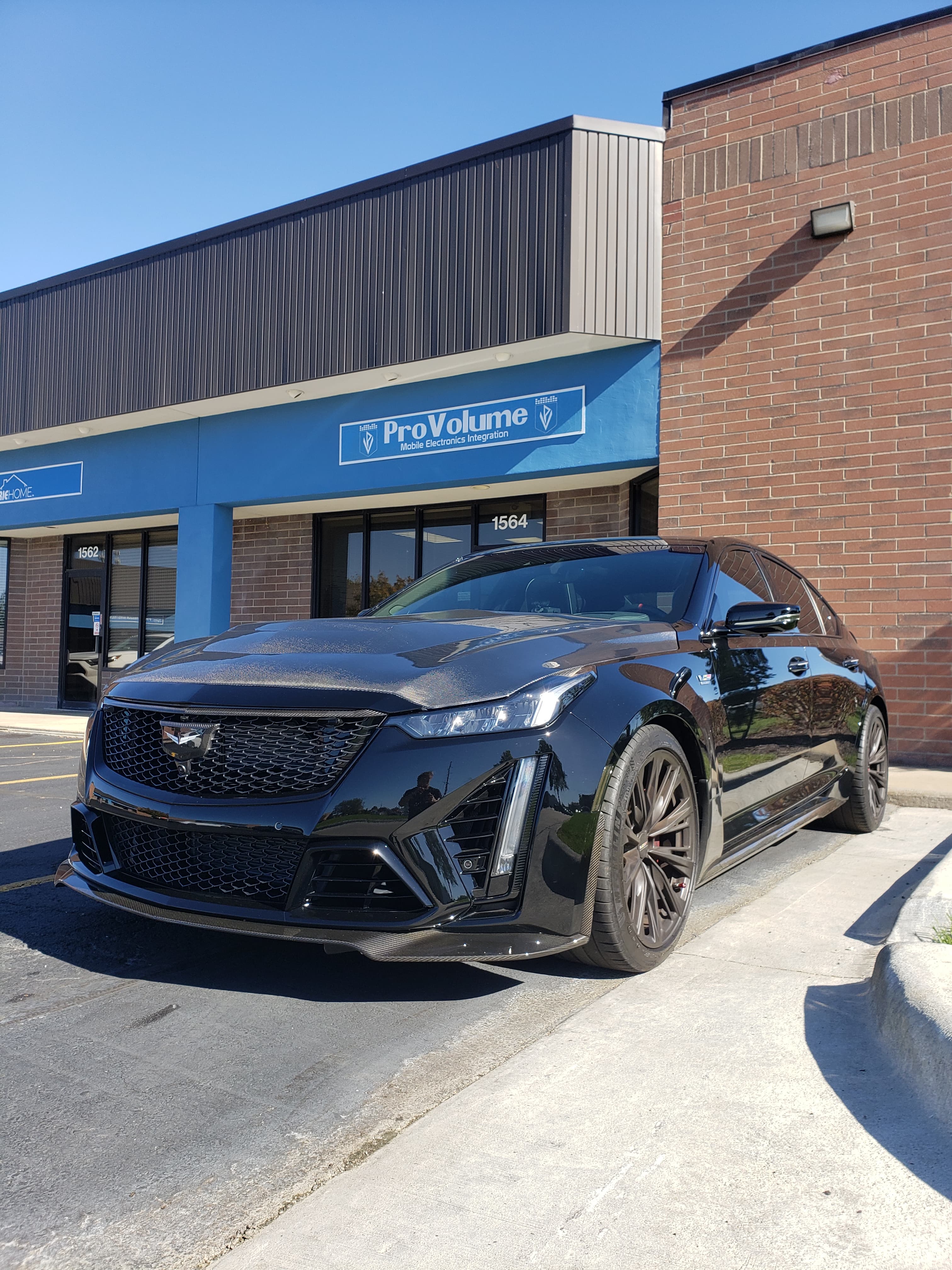 Black Cadillac CT5-V Blackwing with carbon fiber hood parked outside a mobile electronics shop.