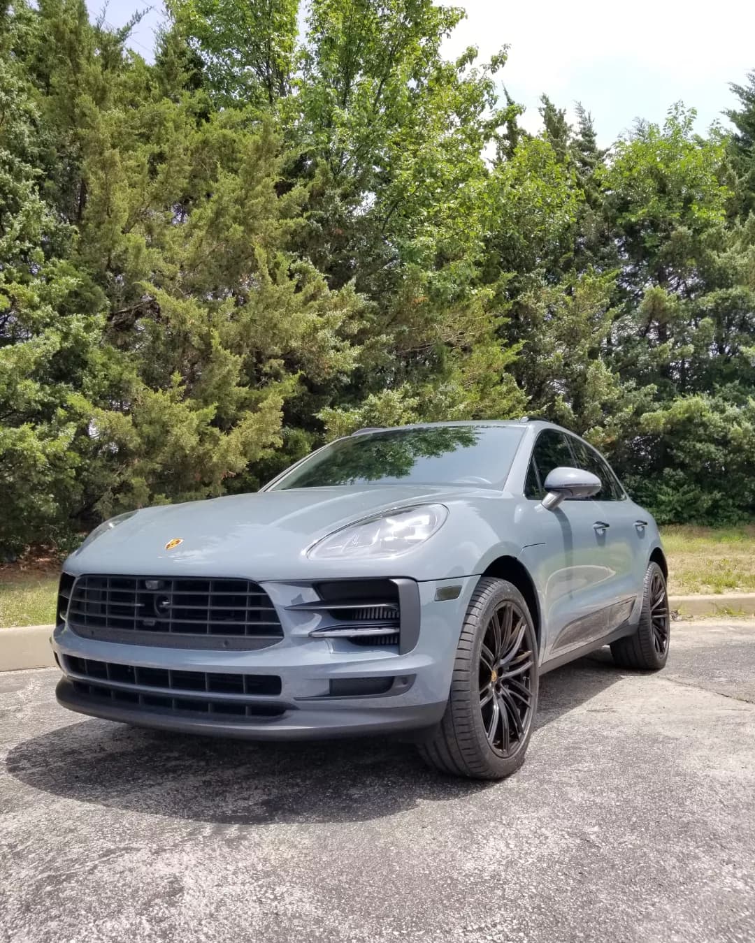 Sleek grey Porsche Macan SUV with black wheels parked on asphalt before a wooded background.