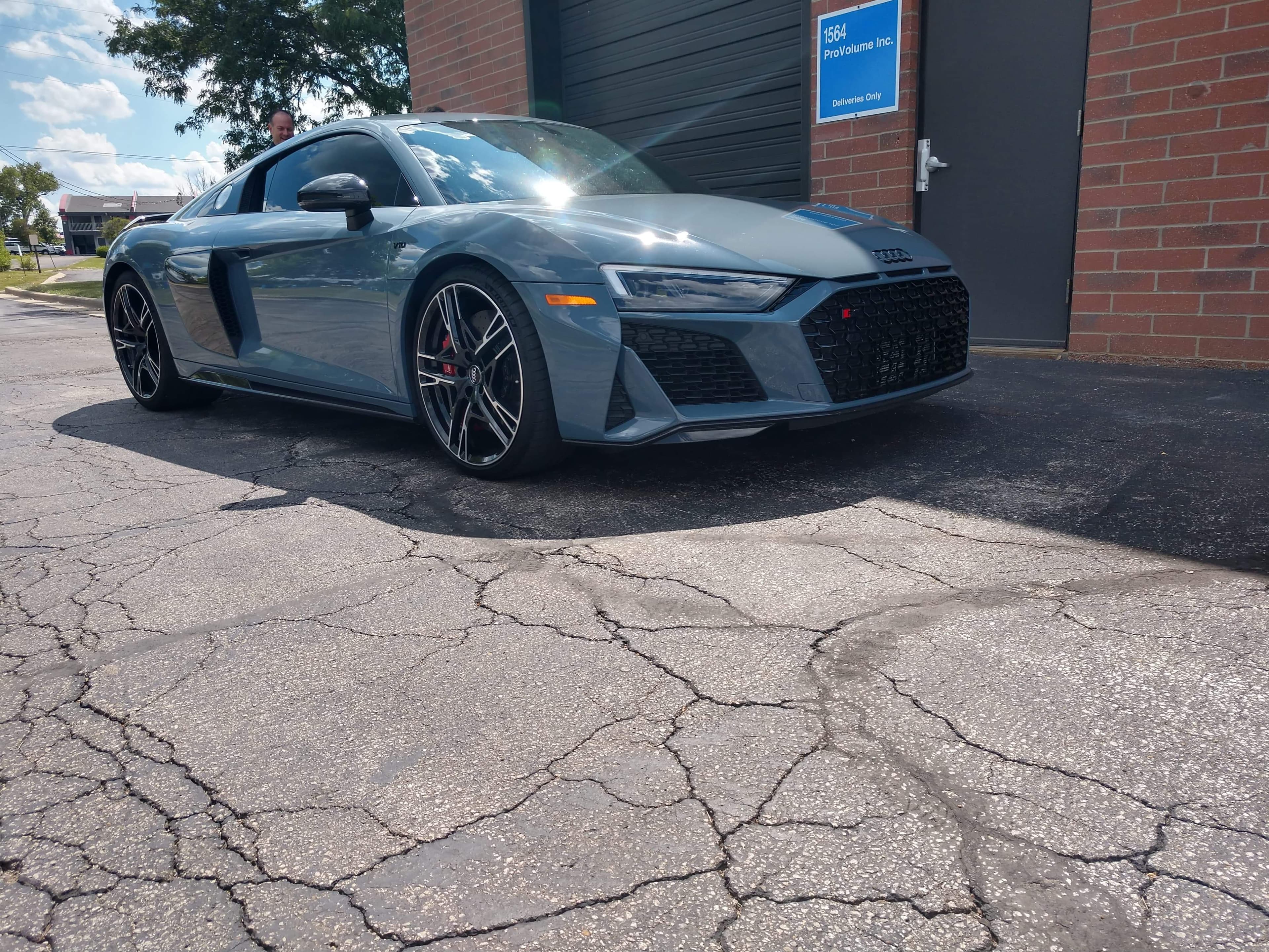 Grey Audi R8 sports car parked on cracked asphalt in front of a brick building.
